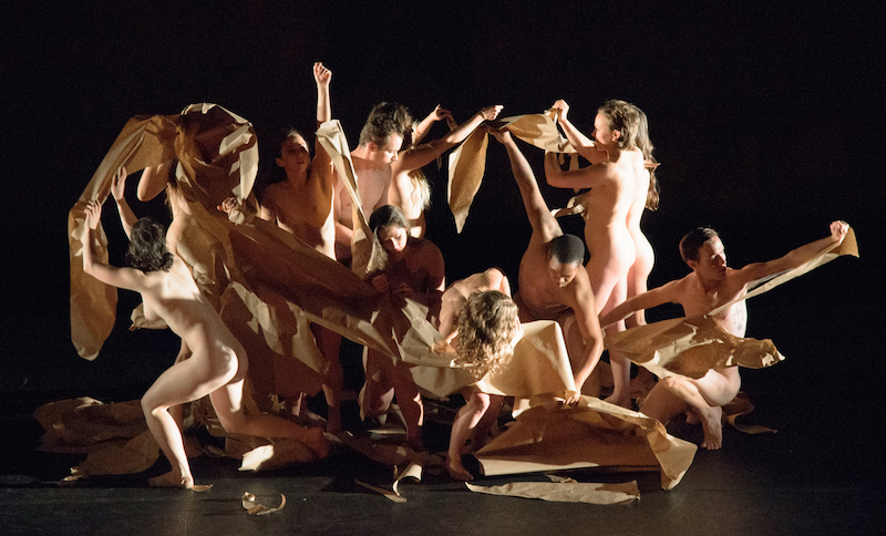 A group of the naked dancers tear paper. The sheets fall and curve around the mass of the people.