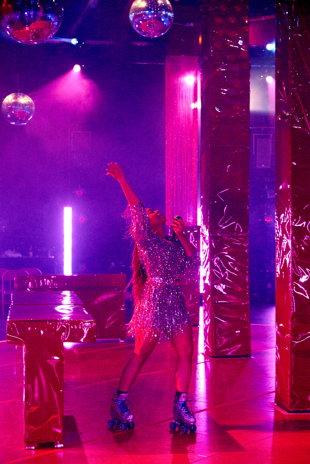 underneath glittering disco balls, a woman in a dripping glittery mini dress wearing silvery roller skates, raises her chest an arms to the cieling while singing into a microphone. It is her club.