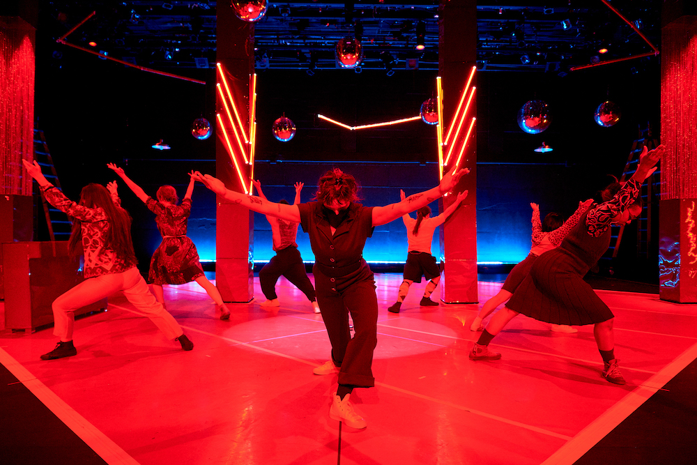 underneath red lighting the seven artists in the cast of Wednesday are positioned in a circle lunging  away from the center with arms outstretched on a high diagonal and their focus to the floor