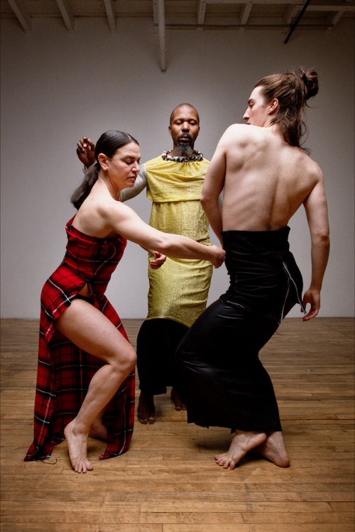 Three dancers in body-hugging long sheath dresses or skirts. One wears a red plaid strapless dress that has a slit up its side. Another wears a gold tunic.