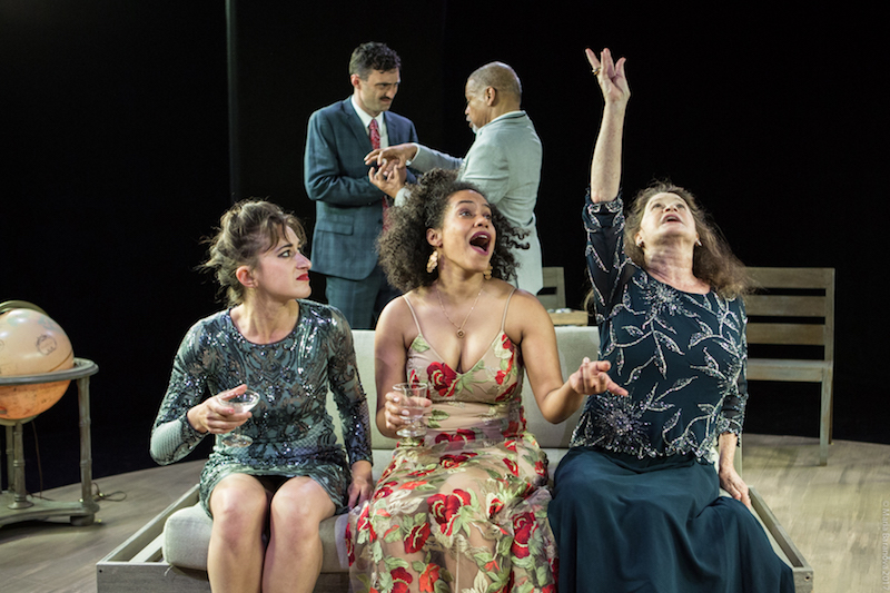 Three women in evening gown sits on a couch and watch one woman gesture her arm high in the air. Two stand in the background speaking intently.