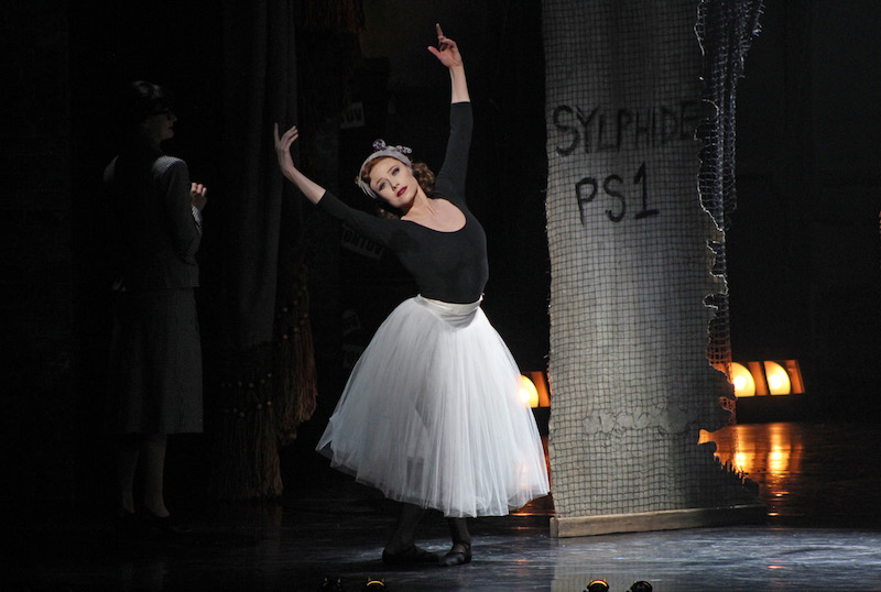 Sara Mearns as Victoria Page wears a black leotard and romantic era white tutu. She stands in a classical ballet post - fifth position with the legs and feet.