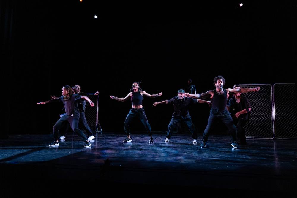 in black outfits against the the black background the company of Rome and Jewels spread their arms to the side in unison seeming to sing