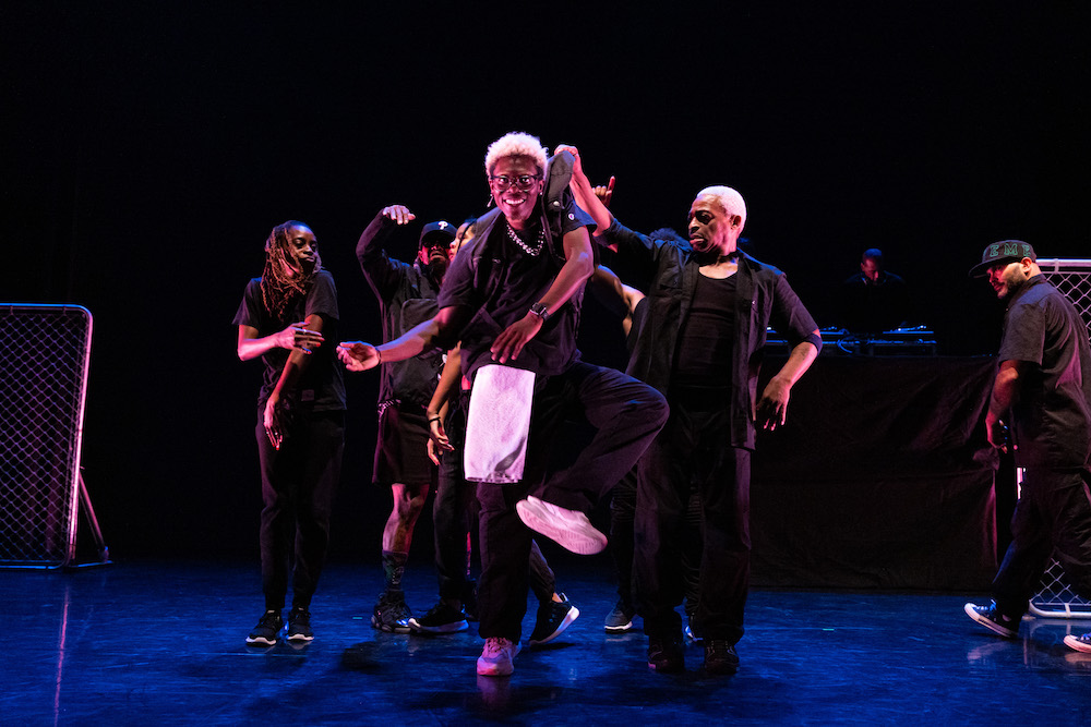 the older narrator lifts a dancer by the back of his shirt as company watches on