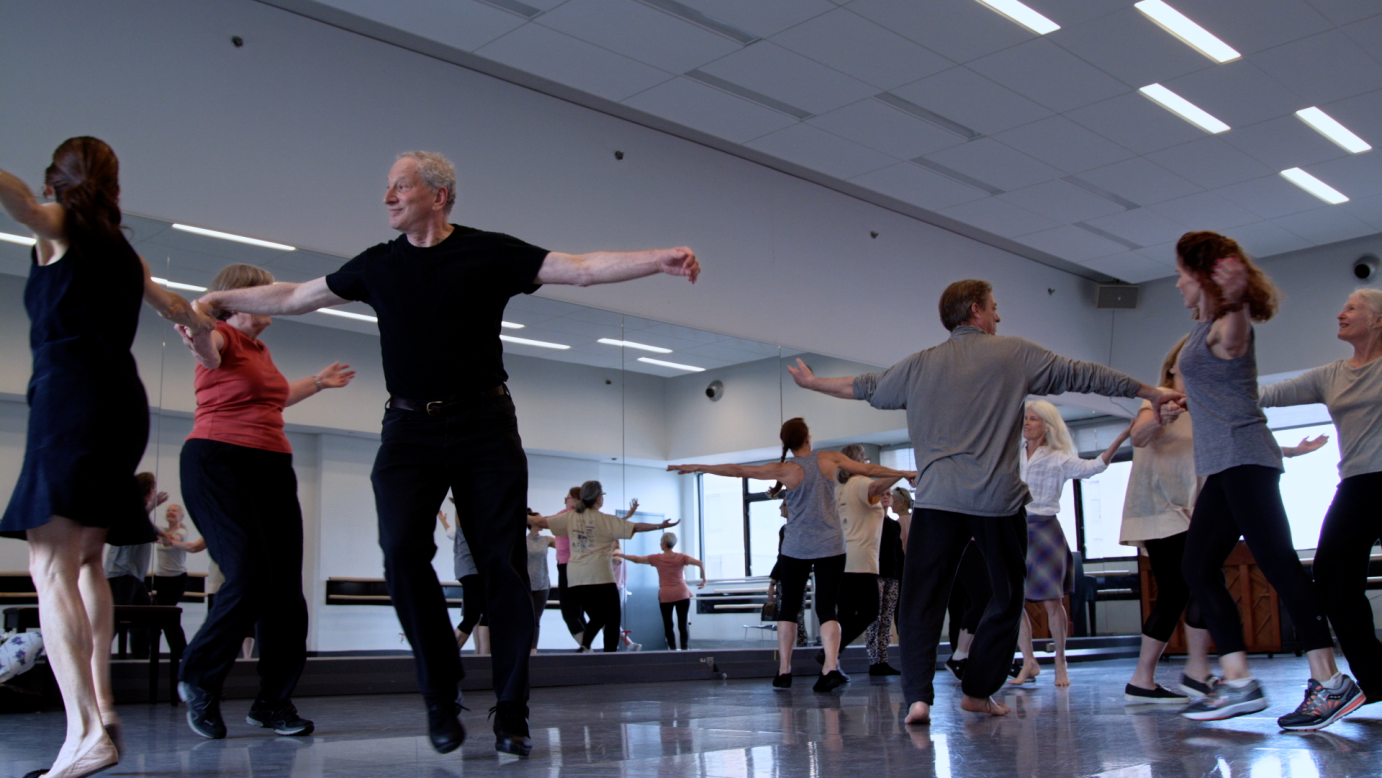 Elder dancers fill a light-filled, grey-floored studio.