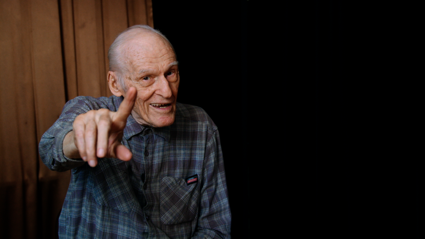 White-haired elderly dancer dressed in a plaid shirt wags a finger while smiling