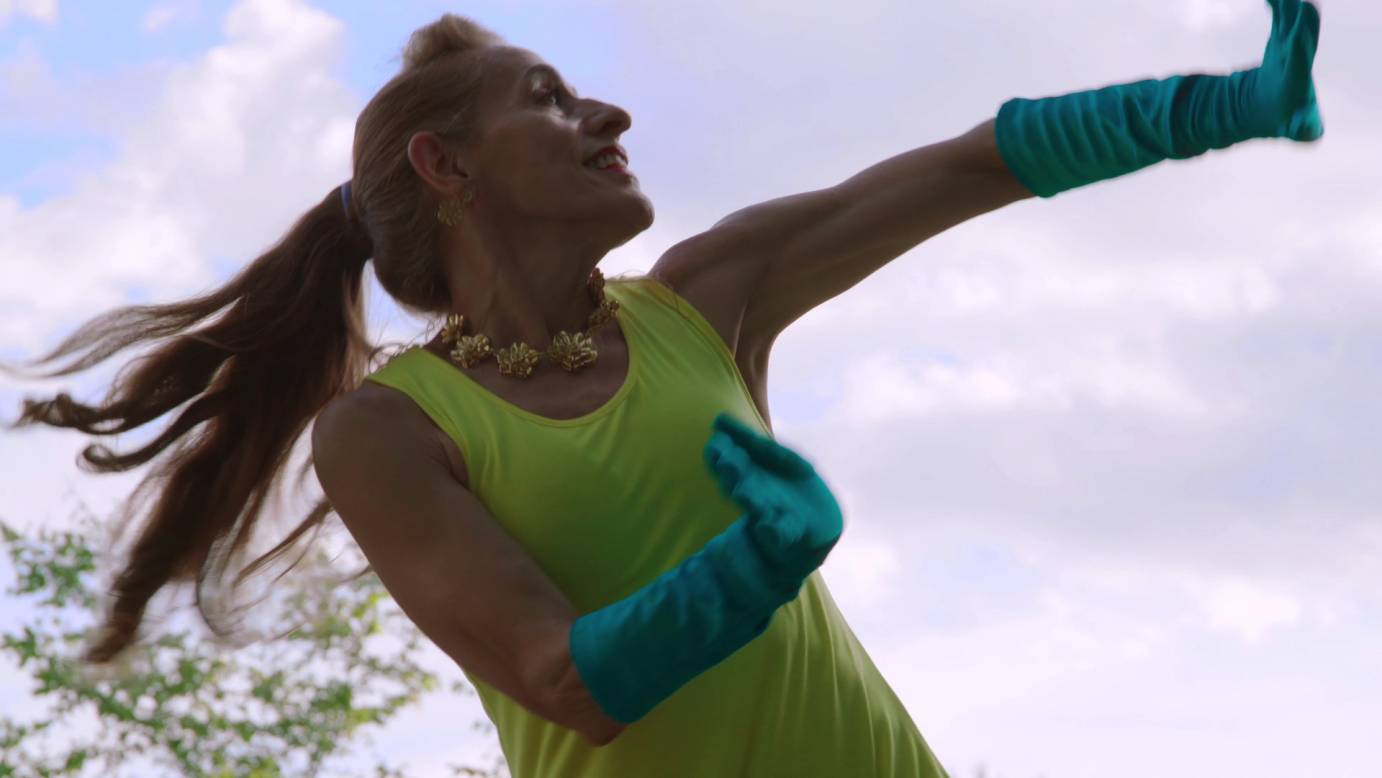 Woman with long ponytail, smiling brightly, wearing a bright yellow sleeveless dress and elbow length blue gloves. A gold necklace of large flowers and dangling gold earrings adorn her. She is dancing against a light blue sky.