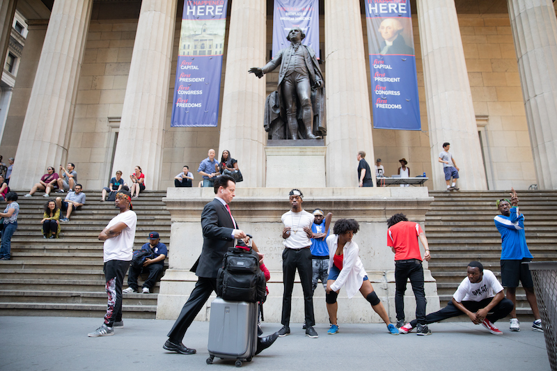 Performers dislocate their shoulders and cross their elbows on Wall St as passersbys walk.