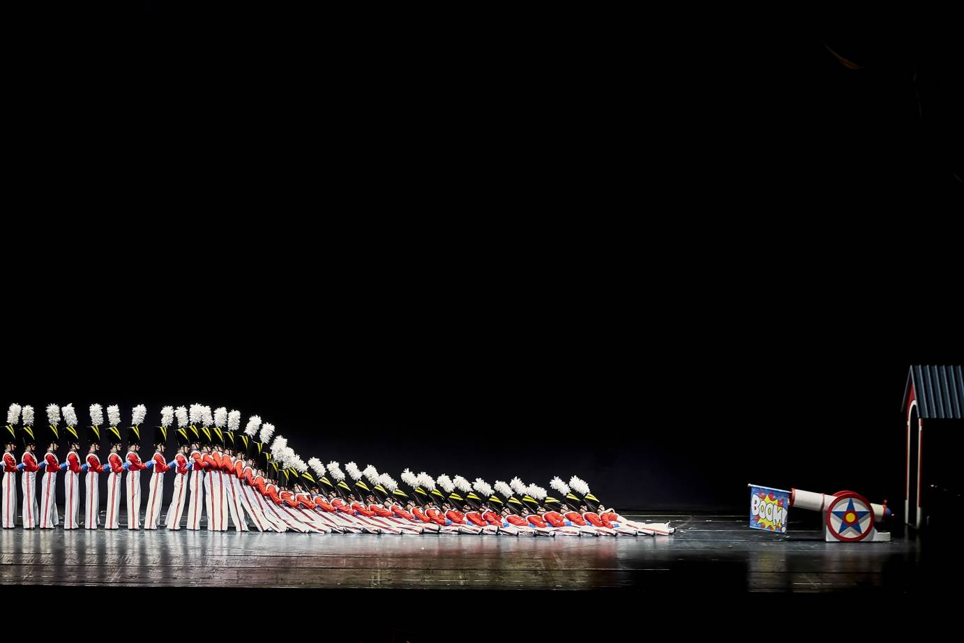 40 Rockettes dressed as fanciful soldiers with white pants that are striped along their sides, w red shirts and white suspender, and tall black hats with plumes. some of them in the back of the line stand upright as the first
