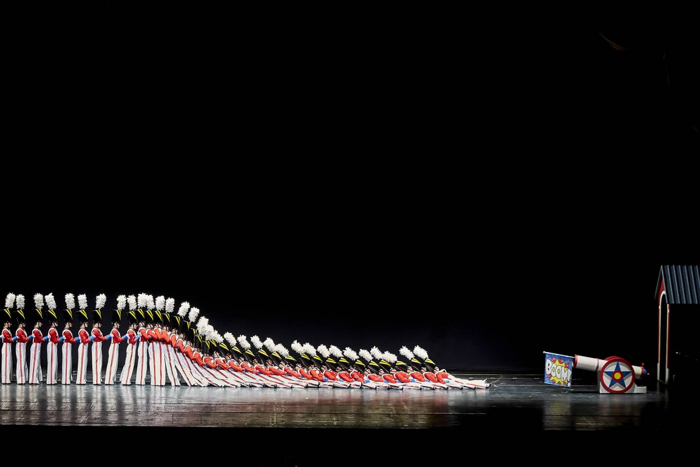 40 Rockettes dressed as fanciful soldiers with white pants that are striped along their sides, w red shirts and white suspender, and tall black hats with plumes. some of them in the back of the line stand upright as the first 