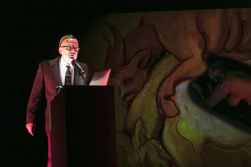 A bespectacled Roy Athey wears a suit and tie and speaks from a podium.