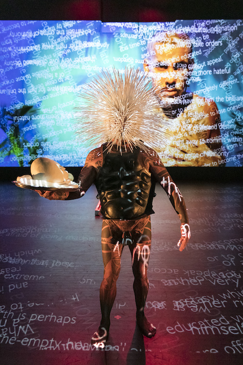 Roy Athey wears what looks like a scuba suit and a spiky headpiece that covers his entire face. He folds a tray of objects. 