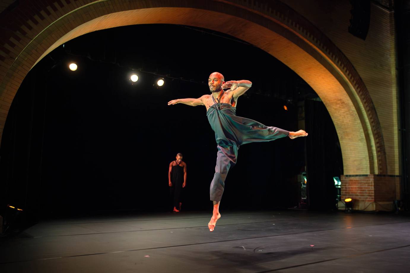 with the arch of Harlem stage creating a striking background, one dancer hovers in the air in the midst of a striking leap in the air. He appears sharp like an arrow flying in space. In the background, under the arch,  another dancer thoughtfully stands staring down at the floor