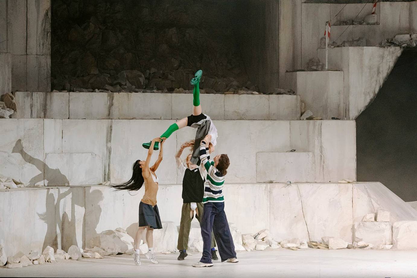 against a magnificent concrete monstrosity a group of dancers in street clothes lift a woman with green boots  upside down in the air... she is being hurled 