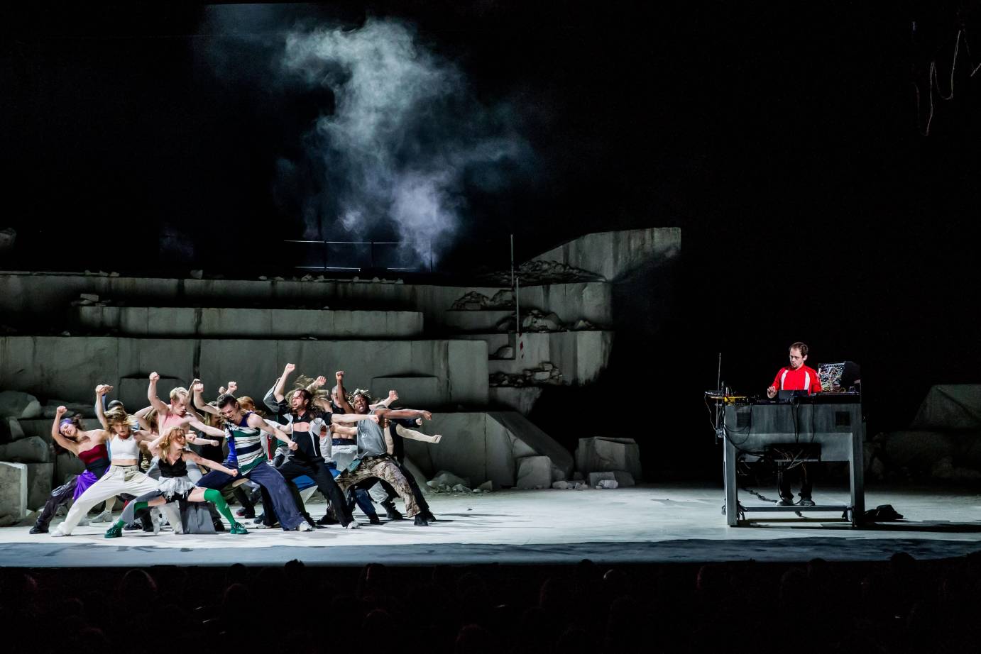 as the DJ Rone wearing a red shirt is off to one side of the stage laying beats down, a group of dancers perhas around twenty in street clothes, lift one fist in the air as if ready to fight, punch or rebel. behind them is a partially decimated concrete structure, smoke rises from the top of the structure