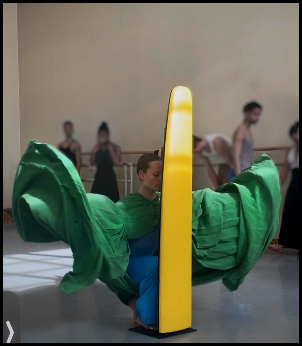 A kneeling dancer tenderly leans her forehead into a standing, thin-line, yellow sculpture. Her upper body is costumed in green, the fabric extending far beyond her horizontal arms. 