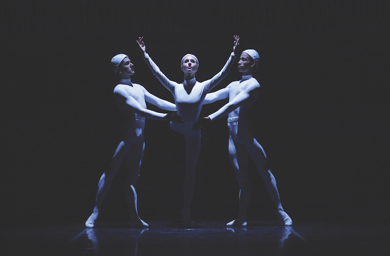 Dancers in unitards that also cover their heads. One dancer balances en pointe with her arms outstretched. Two other dancers stand next to her.