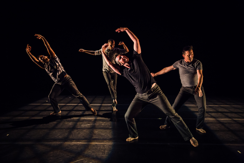 A group of dancers in dark t-shirts and jeans strike various poses. Two have their arms above their heads. Another glances toward his hand which is parallel to the floor.