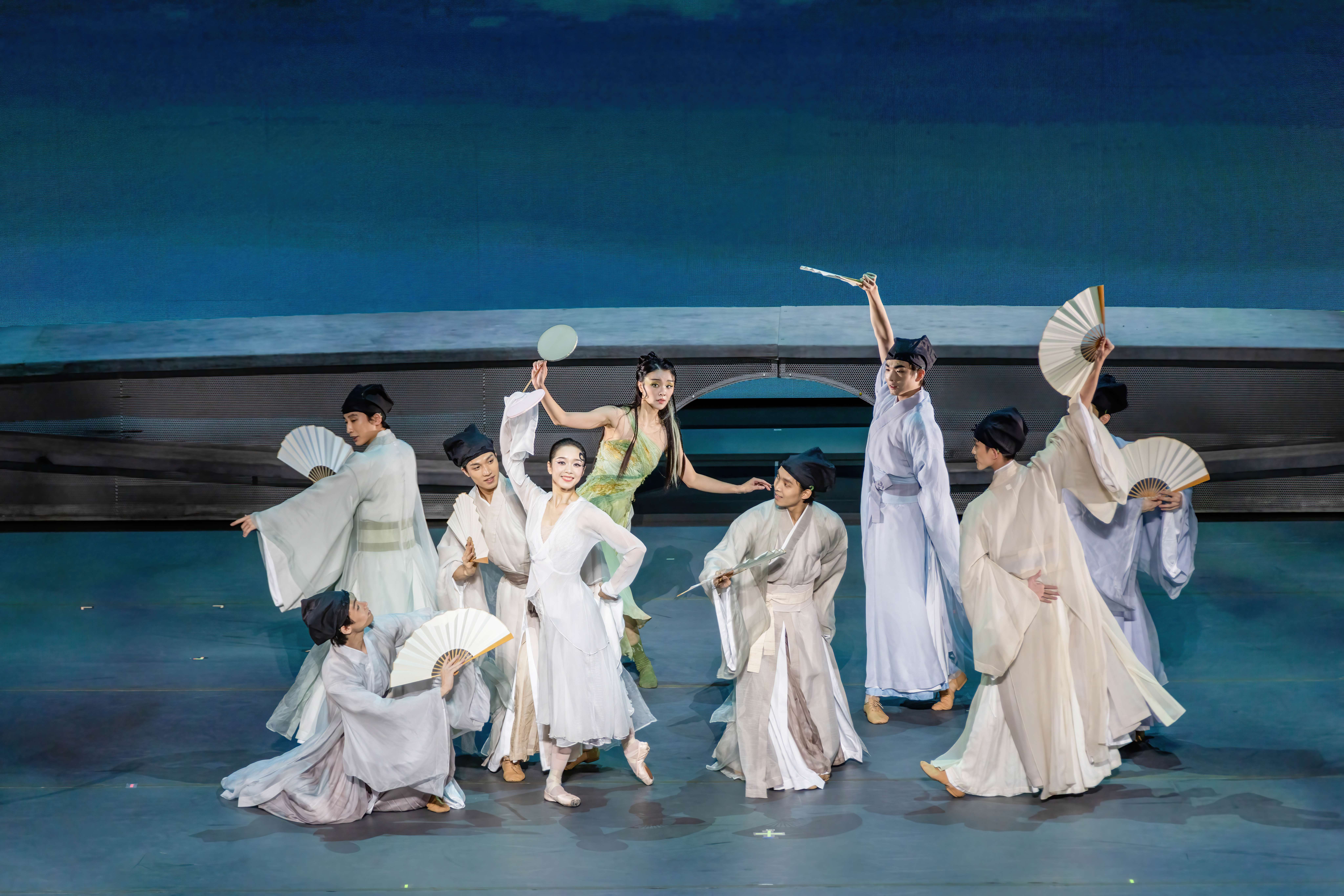 Eight dancers dressed in long, flowing light colored kimonos and dresses, hold fans and are paused in various poses.