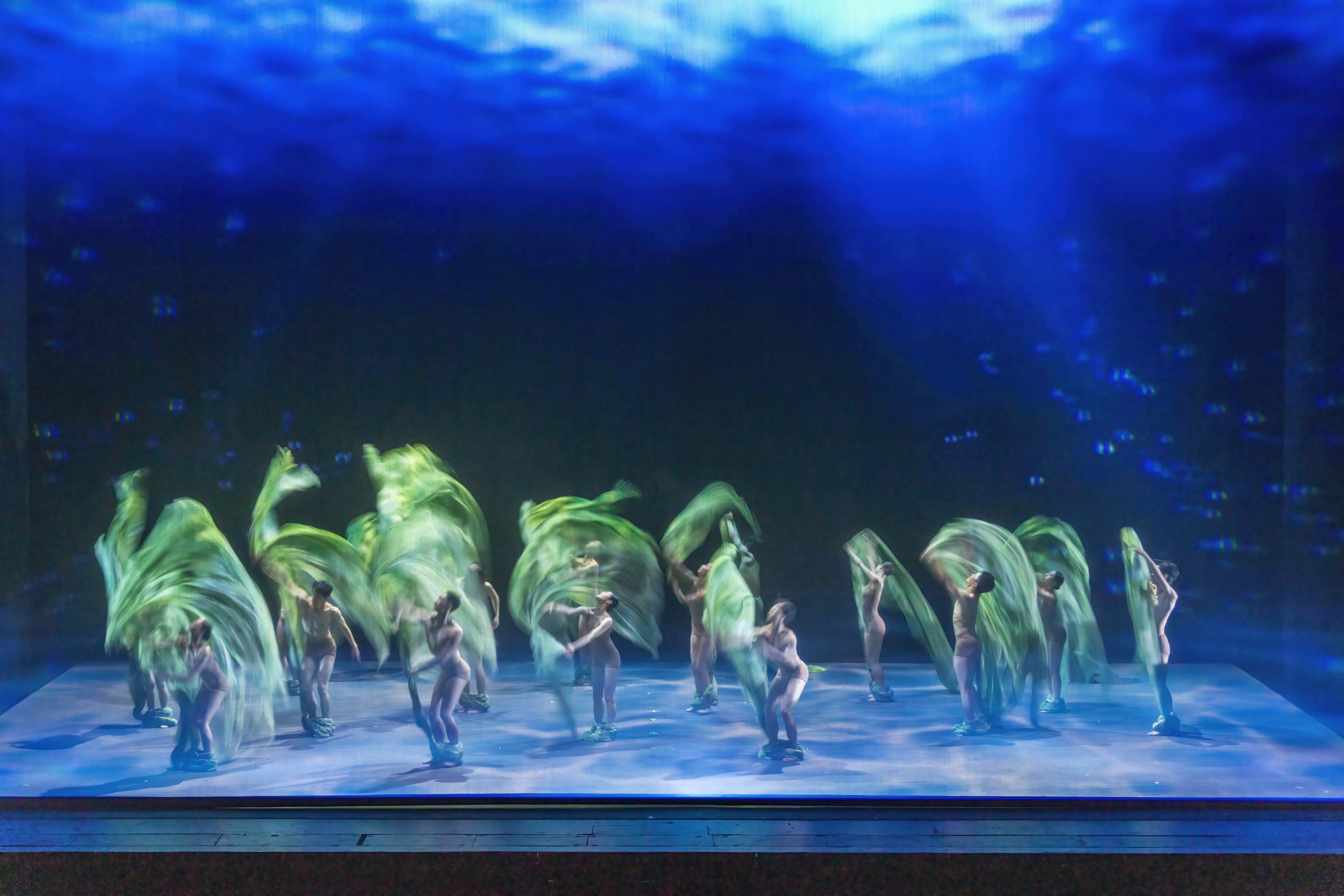 The company swirls green fabric above their heads and around their bodies against a dark blue background and lighter multi-blue hued stage floor.