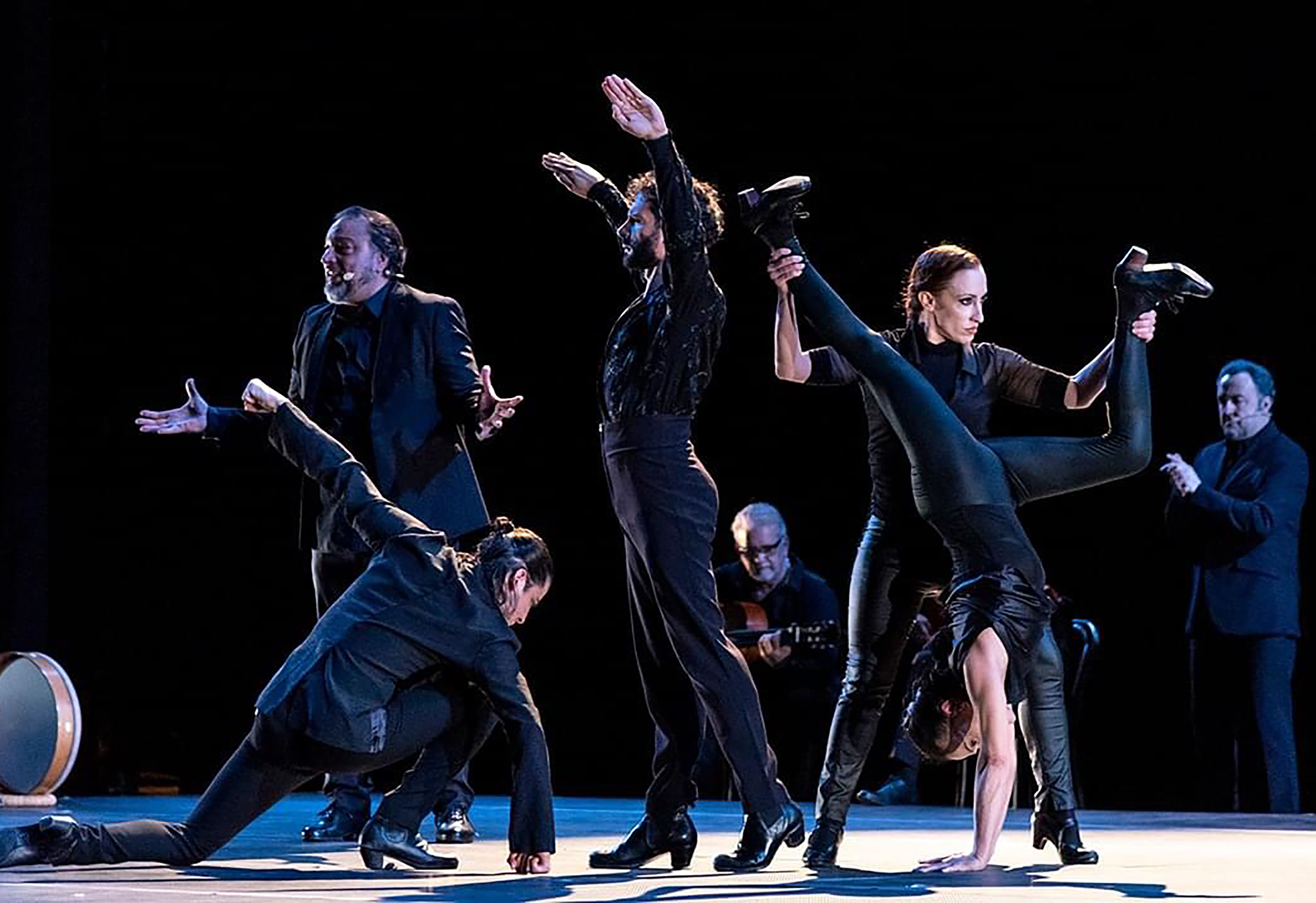 Seven performers including dancers and singers, dressed in black on varying levels: a woman deeply lunging, a bearded man with arms above head, a woman in a split-legged handstand. A man with palms open and and handless mic at mouth, singing.