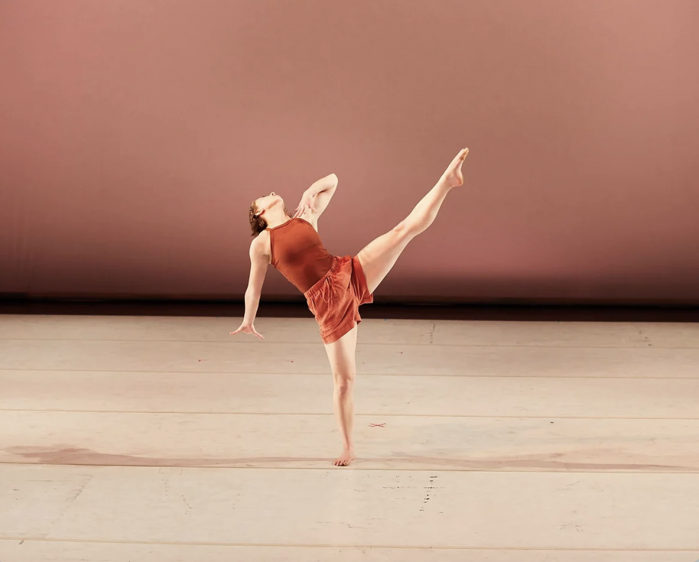 fair skinned woman dancer in rust leotard and lighter rust shorts. Leaning back her face hidden from us, her body faces us as she kicks her left leg far above 90 degrees to her side.