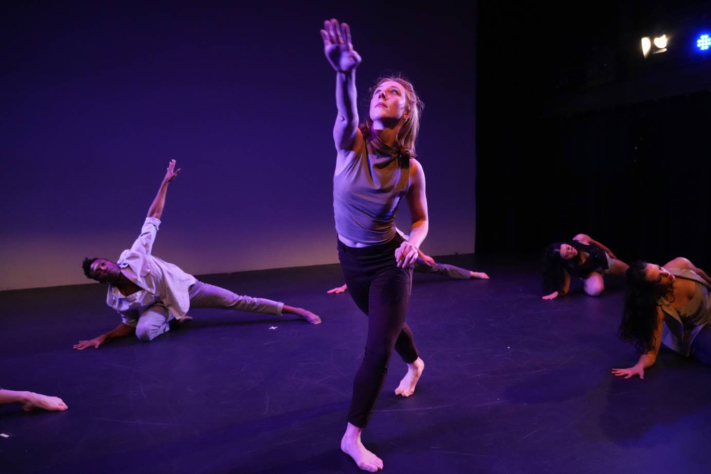 the company in street clothes lie on their sides on the floor as one fair skinned woman stands up and reaches out and upward 