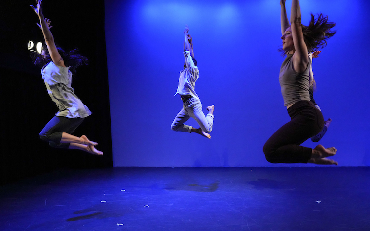 a company of dancers in street clothes jump high above the stage, looking almost as if the are kneeling mid-air 