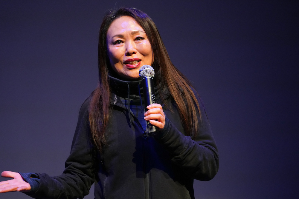 Young Soon Kim at the mic , a Korean woman with long black hair in a warm -up suit talking to the audience