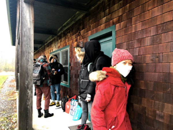 foreground dancer Frances Samson in red and white wool cap with red parka and a white mask covering most of her face on the porch of a rustic wooden inn with green doors there are other people in dark clothes behind her but are anonymous there is luggage on the floor of the porch