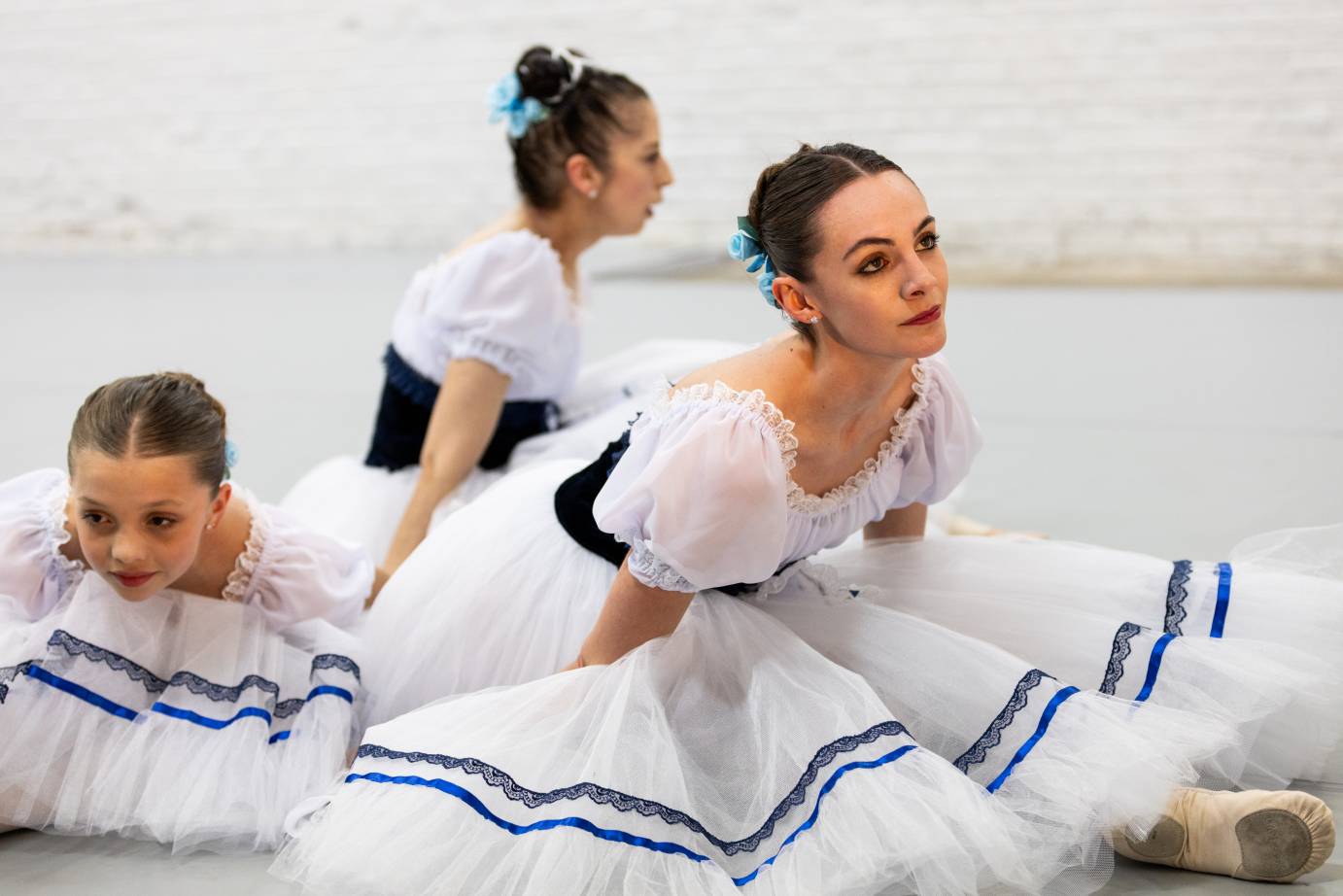 Three ballerinas of different ages: child, mid-20s and veteran are slumped on the floor with their white tulle skirts piled around them.