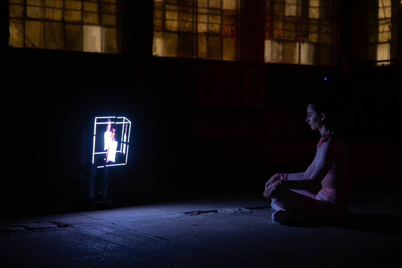 A 3' foot box shaped robot screening images of a talking woman. A pink outfitted woman sits cross-legged in front of the robot with eyes closed.