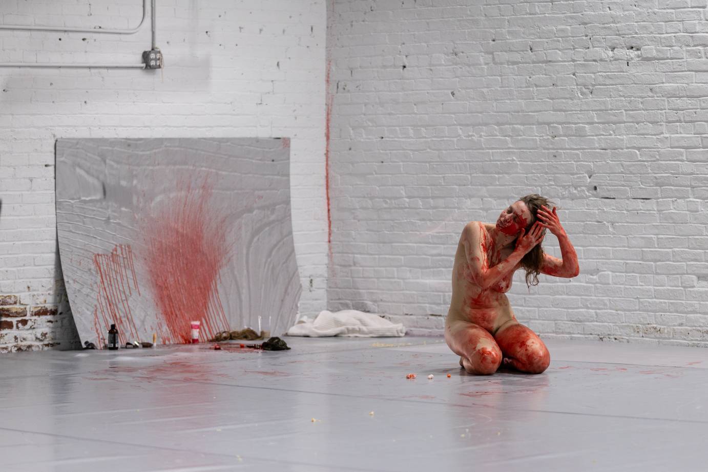 Full-bodied nude woman playing with her dirty-blonde hair extensions. Blood is splattered on the mirror behind her.