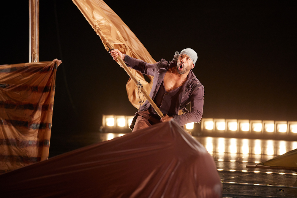 Black man waving what appears to be a mast of a ship -he is dressed in brown rags-barechested and appears to be shouting 