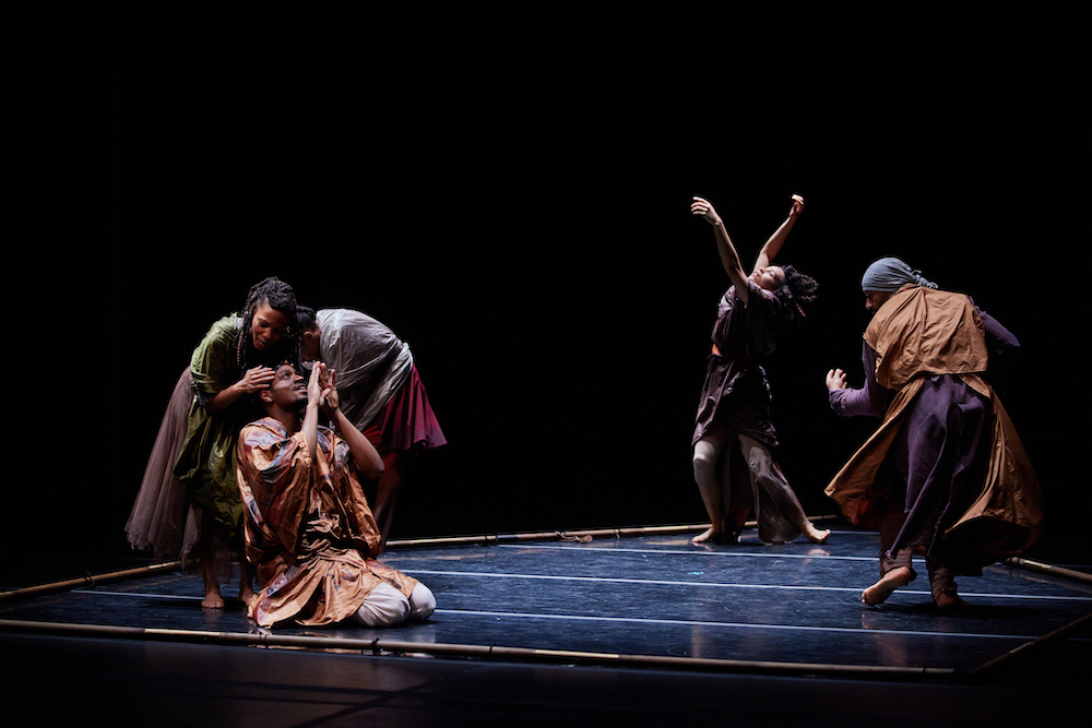 The cast of Embarqued on stage as a trio to the left, a woman standing twisted, knees bent, arms extended to the heavens with her eyes closed, and a figure to the right whose back faces us