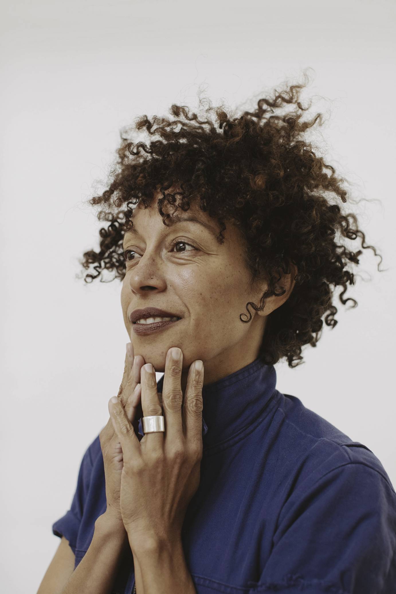 portrait of Stefanie Batten Bland, a light brown woman with curly hair, in a blue top, looking out to the future..her hands touch her chin in thought