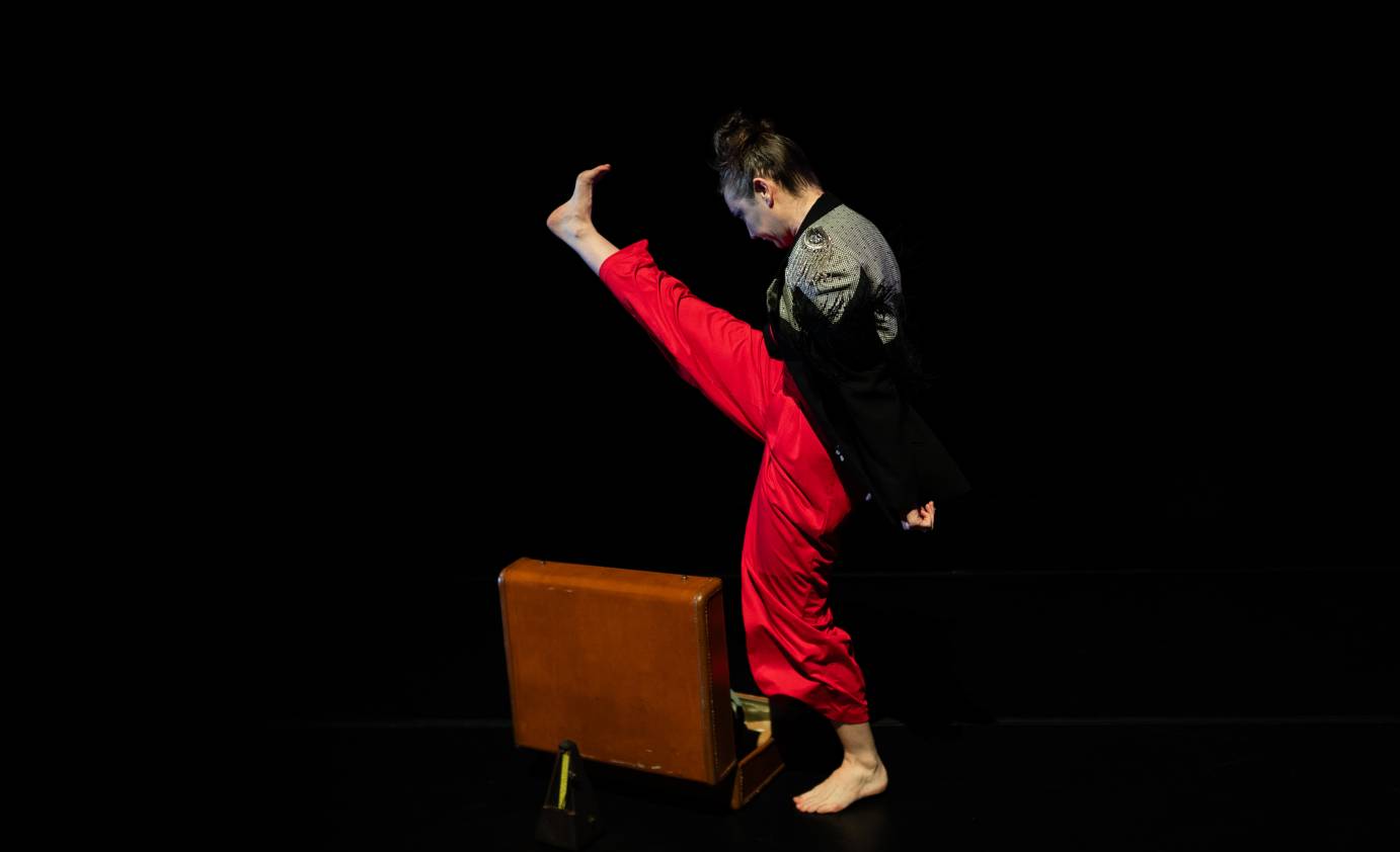 A woman dancer in red pants and black flecked jacket kicks high above a leather suitcase sitting below.