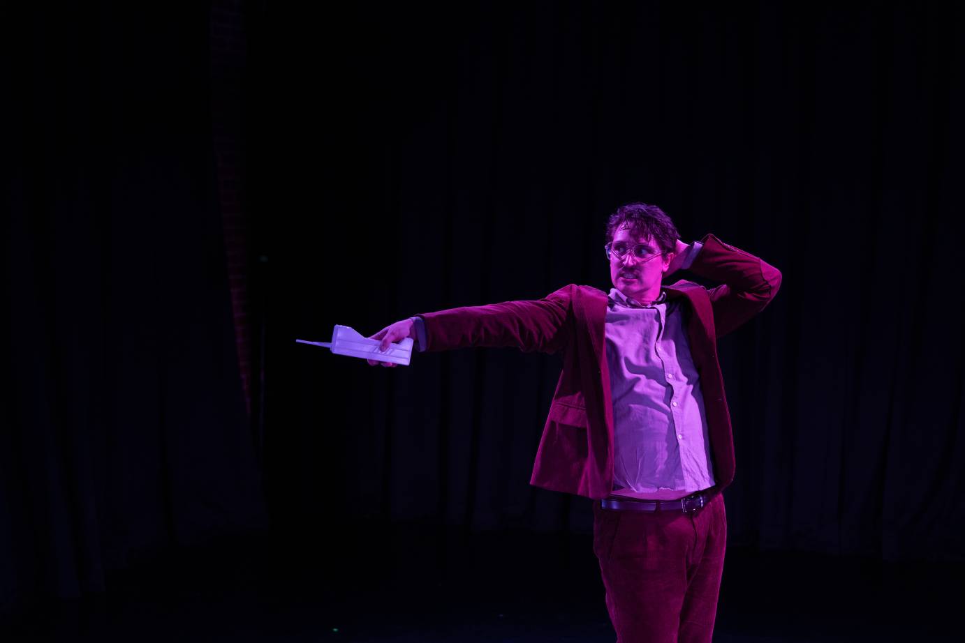 A man in jacket, button-down shirt and pants, touches the back of his head with one hand while the other extends forward holding an old-fashioned phone.