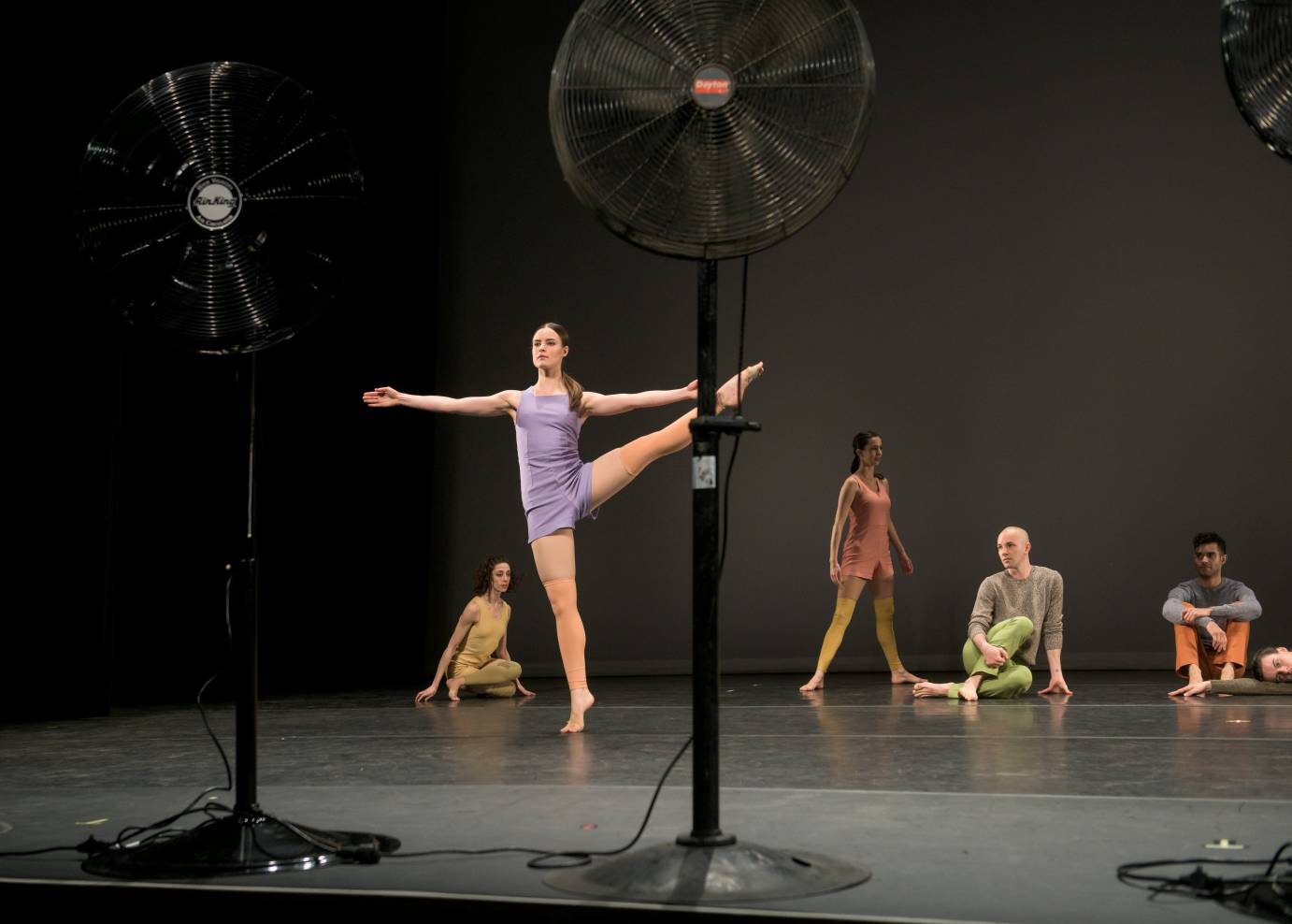 A woman in lilac lifts her leg to the sides as other dancers watch