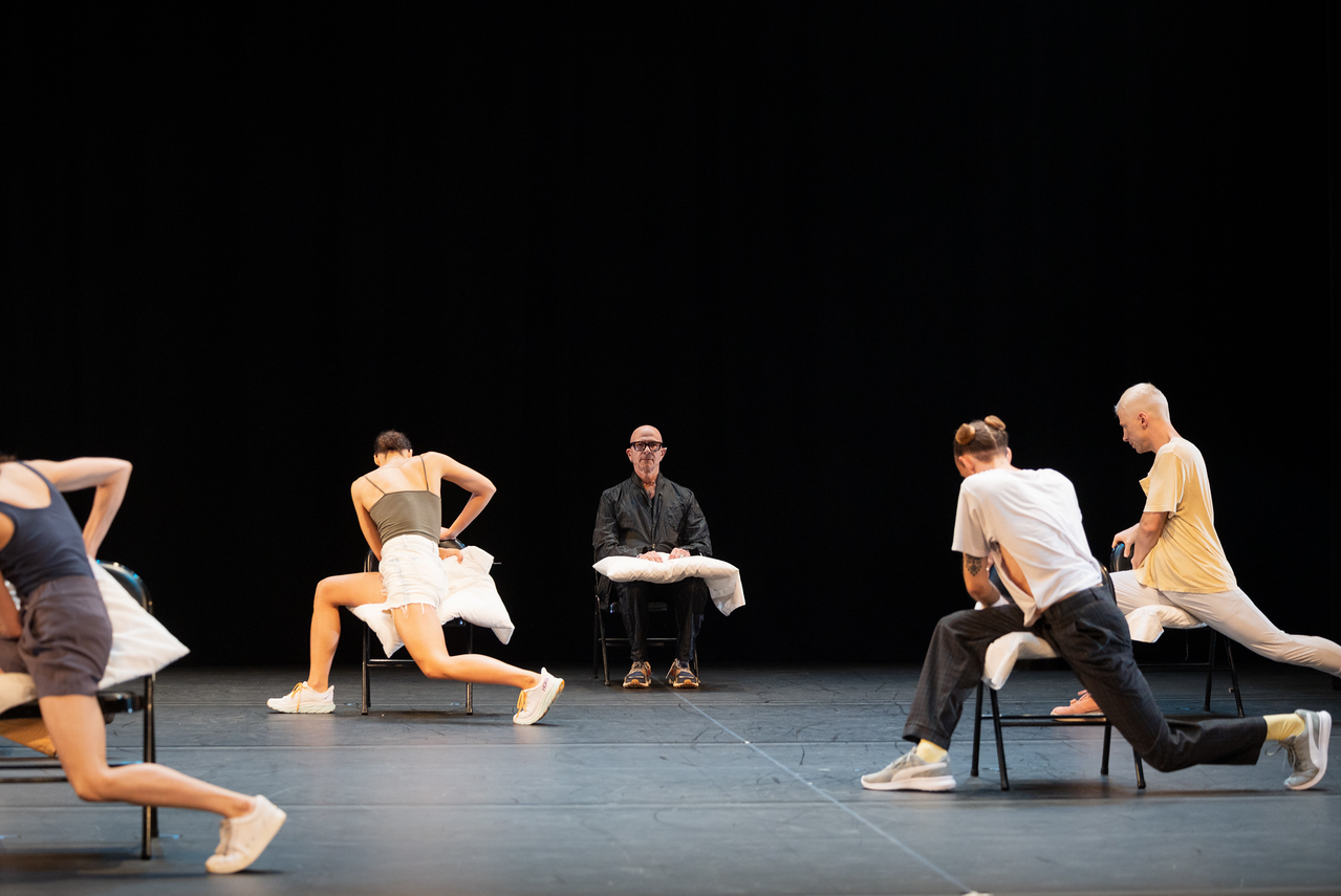 The choreographer sits center stage looking toward the audience flanked by his seated company turned backward on two bent legs with a pillow wrapped on the lower part of their bodies.