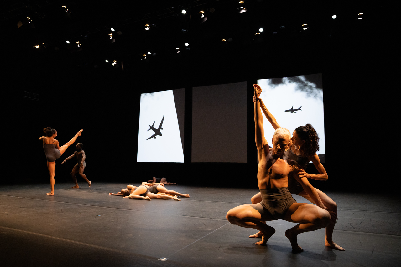 Two black and white background screens showing airplanes in silhouette against which dancers lie on the floor spooning, extend a right leg while standing, press bodies together while deeply plieing. The costumes are gray leotards for women and tight gray shorts reaching halfway up the torso.