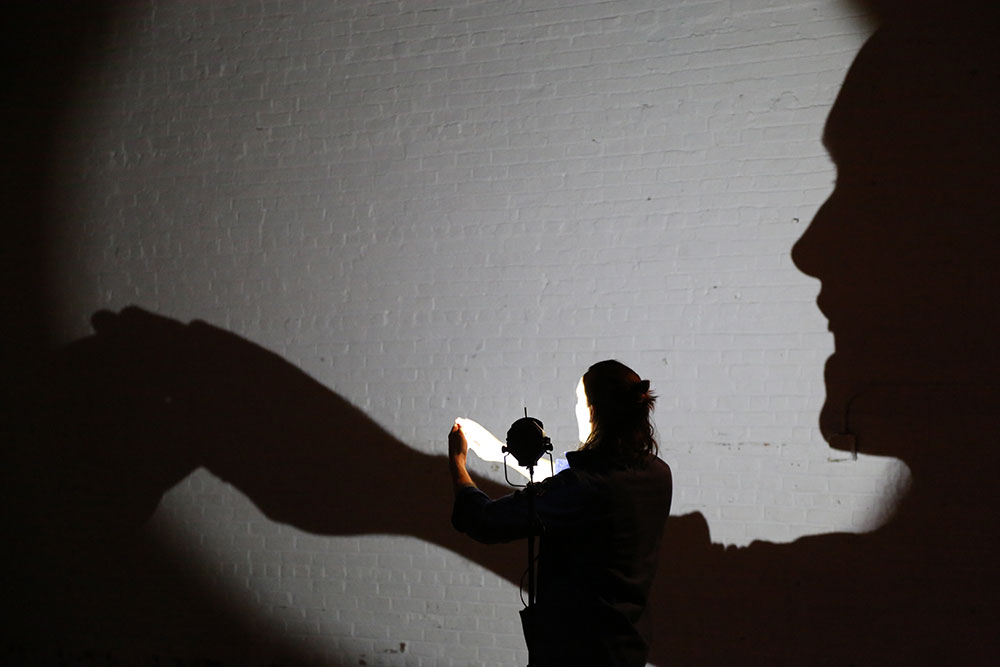 A giant shadow of a human's face and arms are cast on a gray wall. A live performer is at the center of the shot. 