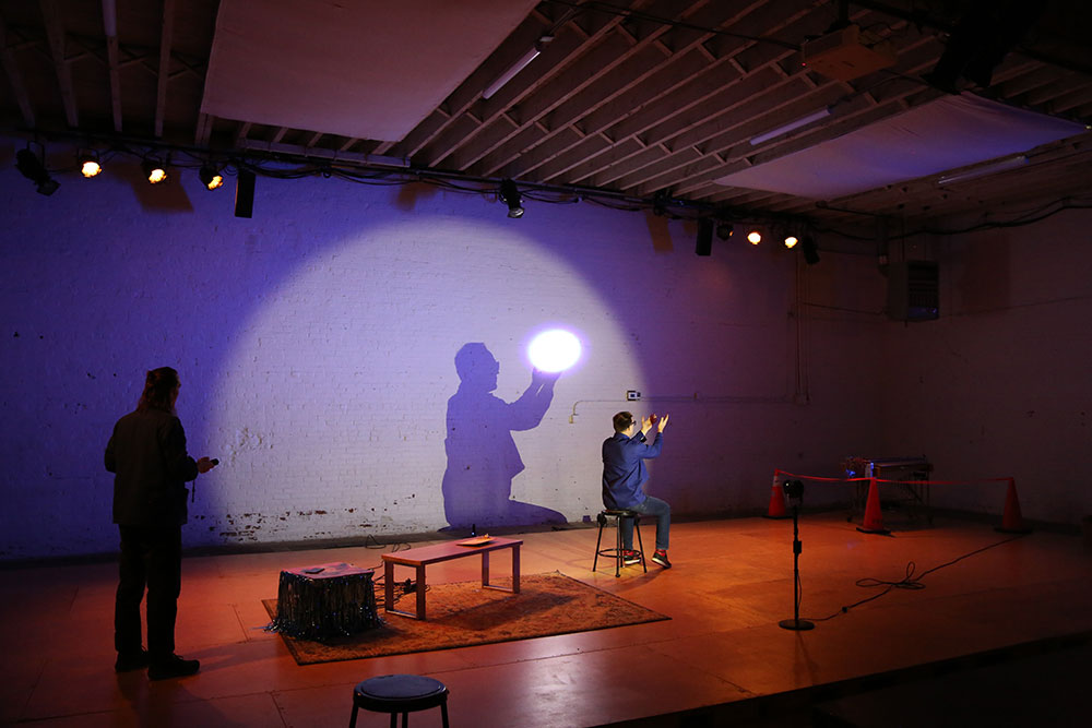 A man sits sideways to the audience with his purple shadow cast on the wall behind.