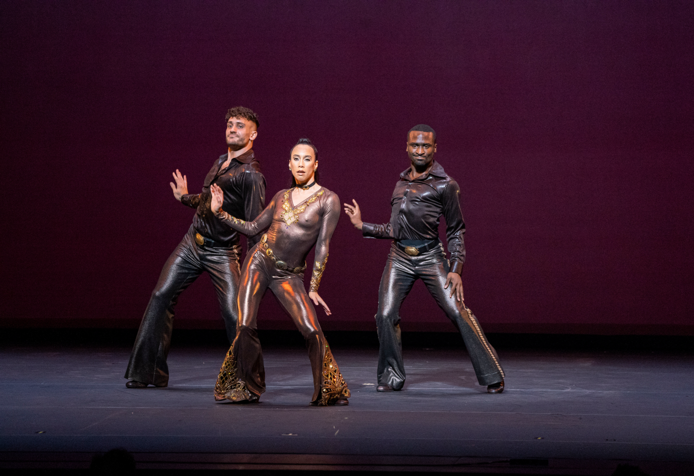 In a bronze, flare-legged unitard stands with two bent legs, one hand up in a relaxed stop sign. Zachary Downer and Tyler Eisenreich make the same pose, flanking her.