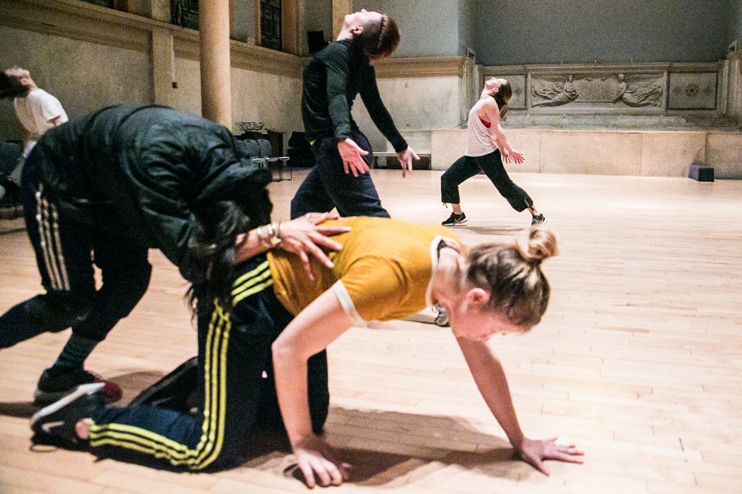 A group of dancers in athletic apparel and sneakers crawl and lunge on Danspace's wooden floor