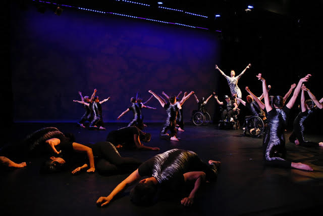 A massive group of dancers kneel and extend their arms in a V as they lift someone in white upward