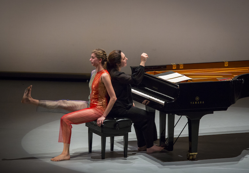 Dinnerstein and Jones sit back to back on the piano bench. Dinnerstein lifts her hand off the piano as if she just finished playing the last note of the variation. Jones flexes her right foot and lifts her leg parallel to the ground.