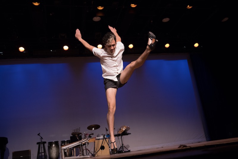 Caleb Teicher swings his arms behind him while his left leg swings out in front. He wears t-strap heeled taps.