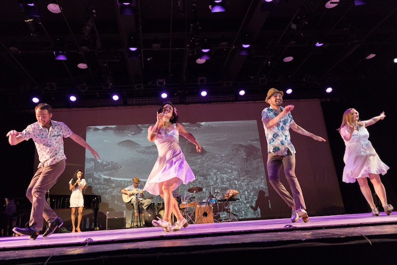 Four tappers in printed shirts, for the men, and dresses, for the ladies. A band plays behind them.