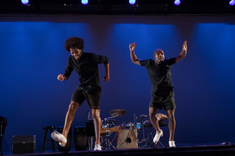 Two men in black button ups and shorts and white taps dance in 